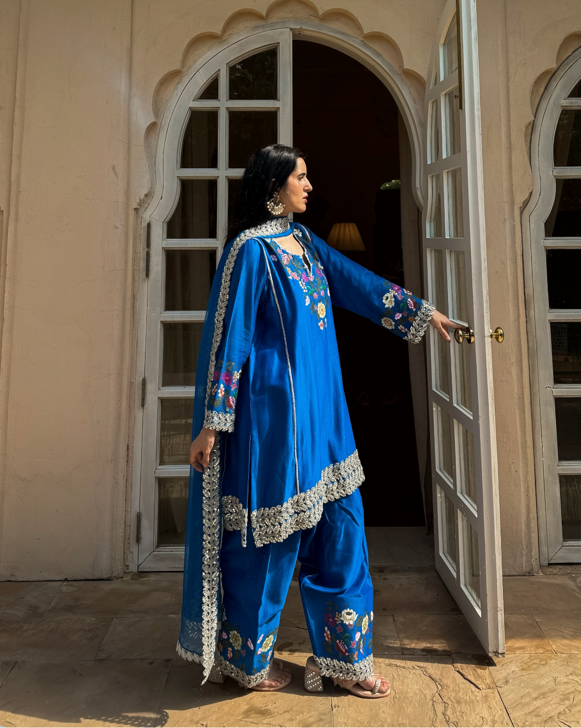 A woman wearing a royal blue embroidered traditional outfit with matching pants and a draped scarf stands in front of an open white door of a beige building, holding the door handle with one hand.
