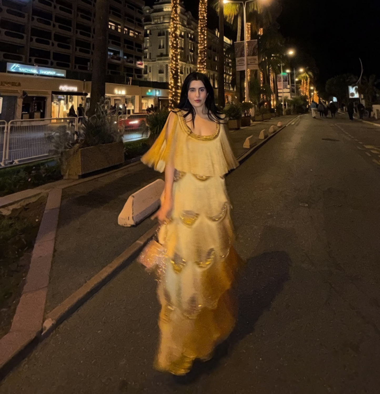 A woman wearing a long, tiered yellow dress walks down a city street at night. The dress has a sheer, layered design with ruffled sleeves and a flowing skirt.
