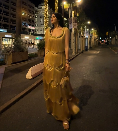 A woman wearing a sleeveless gold maxi dress with gold embroidery walks down a city street at night.