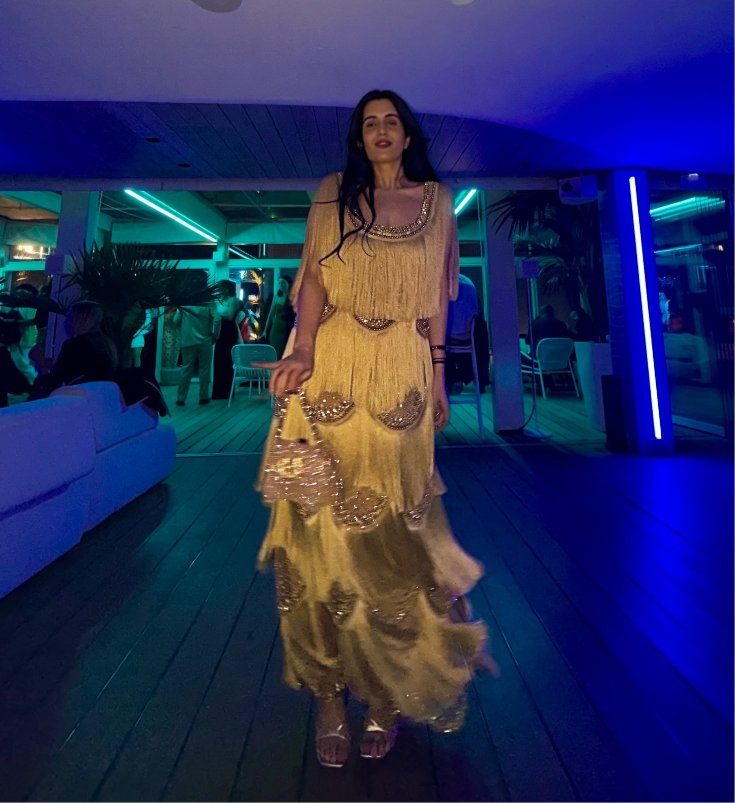 A woman walks down a wooden hallway wearing a long, tiered, gold embellished dress in a dimly lit venue with blue ambient lighting.