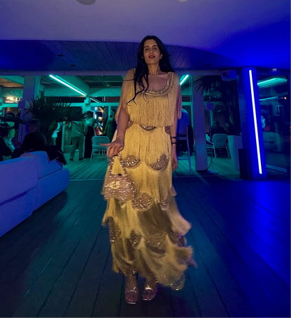 A woman walks down a wooden hallway wearing a long, tiered, gold embellished dress in a dimly lit venue with blue ambient lighting.