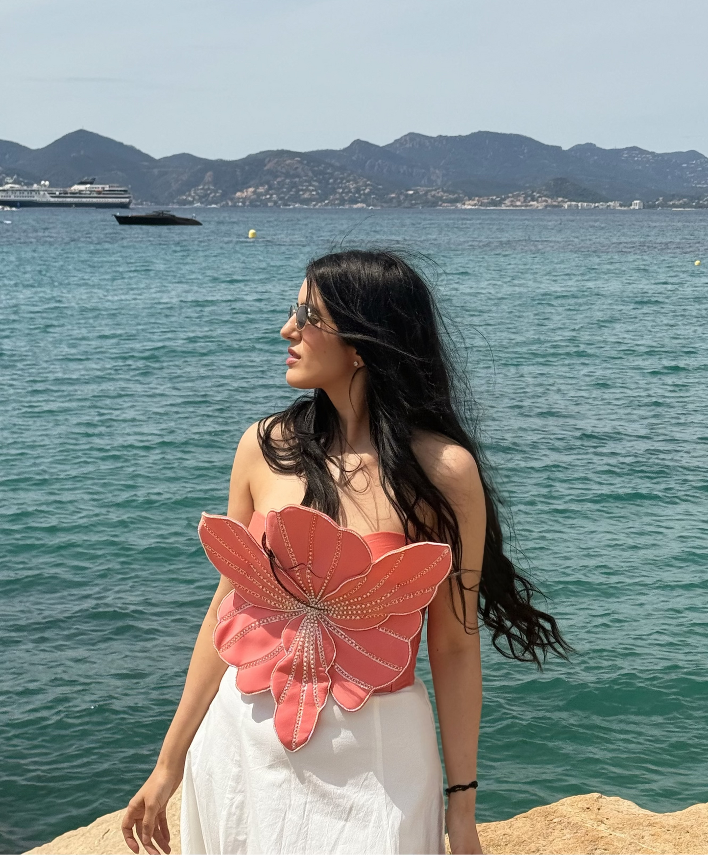 A woman stands on a rocky shoreline by the sea, wearing a white strapless dress with a large pink flower applique on the front. The background features a turquoise ocean, distant mountains, and a few boats under a light blue sky.