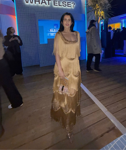 A woman wearing a long, flowing champagne-gold dress stands on a wooden deck at a nighttime event. Behind her is a blue-lit structure with the words "WHAT ELSE?" and a glowing "N" sign, with other guests visible in the background.