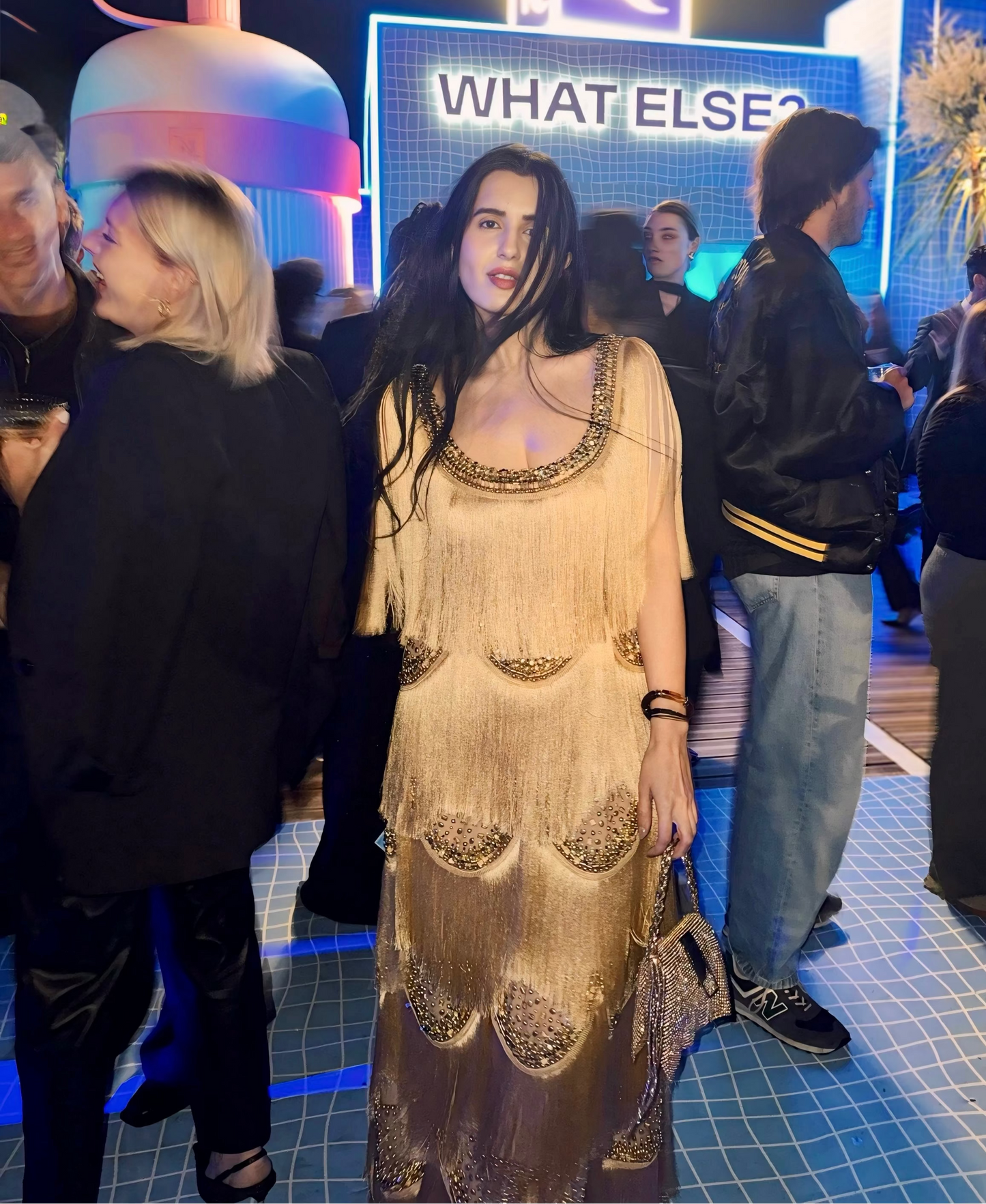 A woman wearing a beige fringed maxi dress with gold-toned embellishments stands in a crowded event space at night. A large illuminated sign behind her reads 'WHAT ELSE?' with a stylized logo above it, and the scene is lit with blue and pink event lighting.