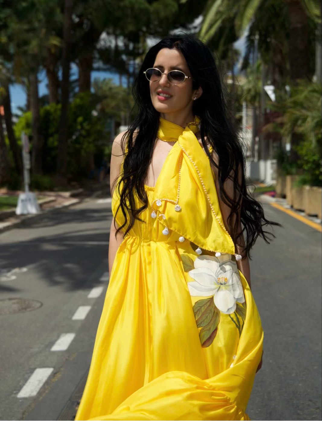 A woman wearing a bright yellow halter-neck dress with a large white flower applique and pearl-like embellishments, standing on a sunlit street lined with palm trees.