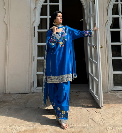 A woman wearing a royal blue embroidered traditional outfit stands in front of an open white arched doorway, holding the doorframe with one hand and gesturing with the other.