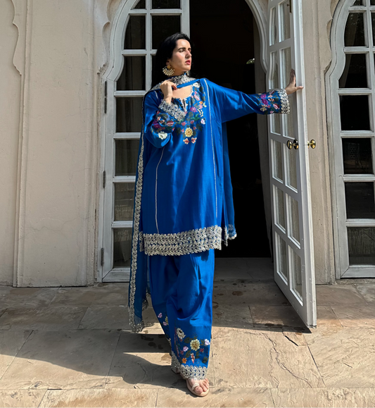 A woman wearing a royal blue embroidered traditional outfit stands in front of an open white arched doorway, holding the doorframe with one hand and gesturing with the other.