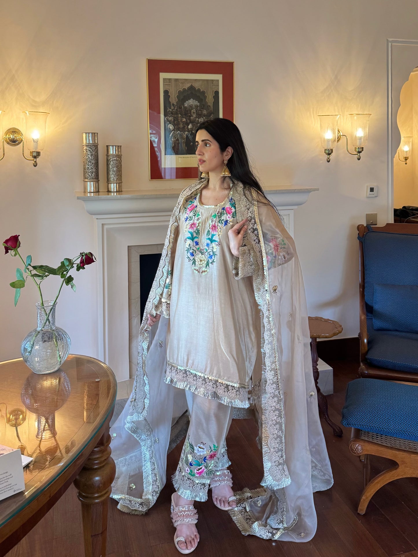 A woman models a traditional South Asian outfit in a furnished room with a fireplace. The outfit consists of a light beige or off-white kurta and matching pants with colorful floral embroidery, paired with a sheer white dupatta featuring silver mirror work and floral embroidery.