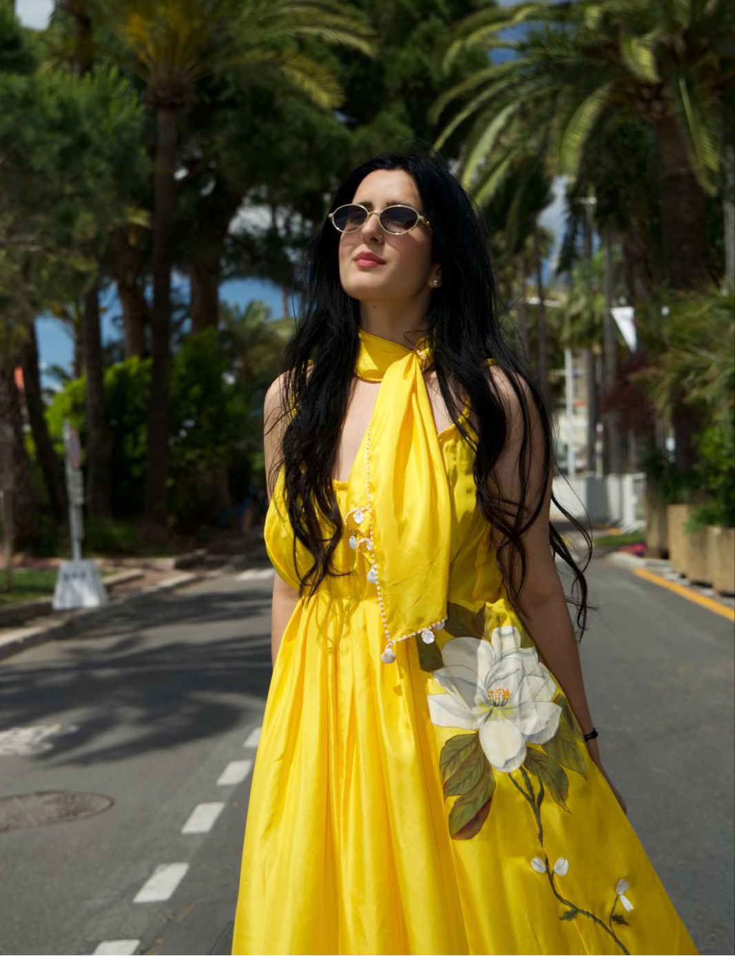 A woman wearing a bright yellow halter-neck dress with white floral embroidery stands on a palm-tree-lined street, wearing round sunglasses.