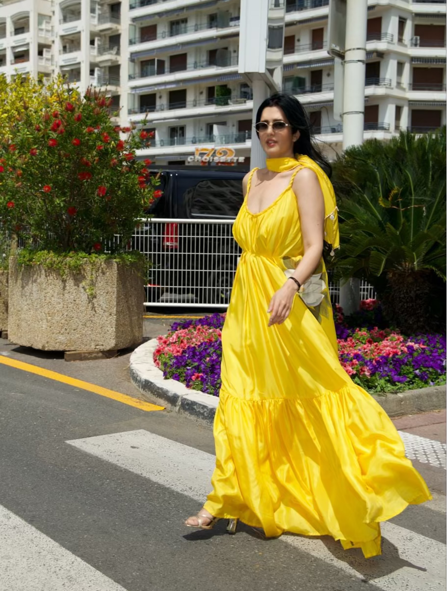 A woman wearing a bright yellow tiered maxi dress and sunglasses walks across a crosswalk on a sunny city street with apartment buildings and flower beds in the background.