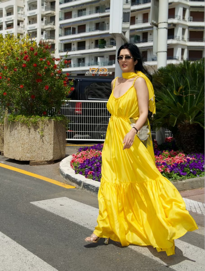 A woman wearing a bright yellow tiered maxi dress and sunglasses walks across a crosswalk on a sunny city street with apartment buildings and flower beds in the background.