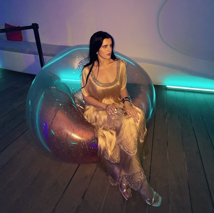 A woman in a shimmering champagne gold gown sits inside a large transparent bubble chair on a wooden floor, with blue and purple ambient lighting in a modern room.