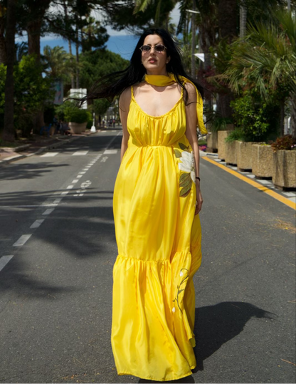 A woman wearing a bright yellow sleeveless maxi dress and matching choker stands on a sunlit road lined with palm trees.