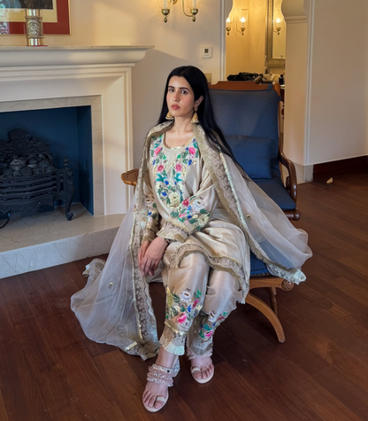 A woman wearing a traditional embroidered outfit sits on a blue armchair in a furnished room with a fireplace, wooden floor, and mirror.