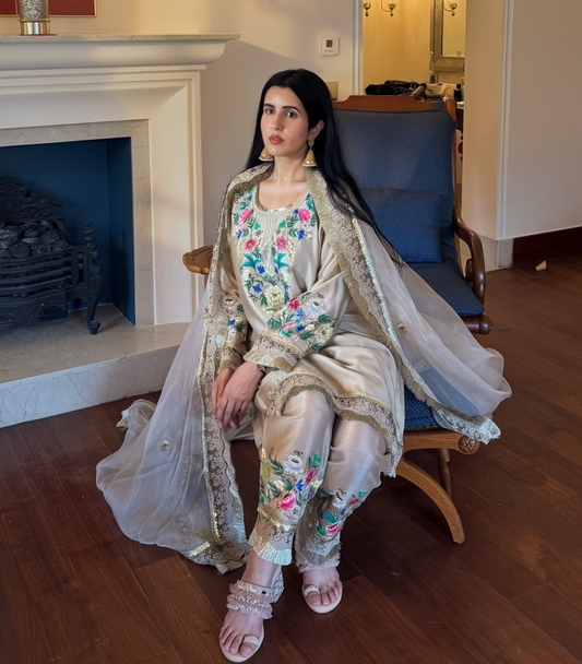 A woman wearing a traditional embroidered outfit sits on a blue armchair in a furnished room with a fireplace, wooden floor, and mirror.
