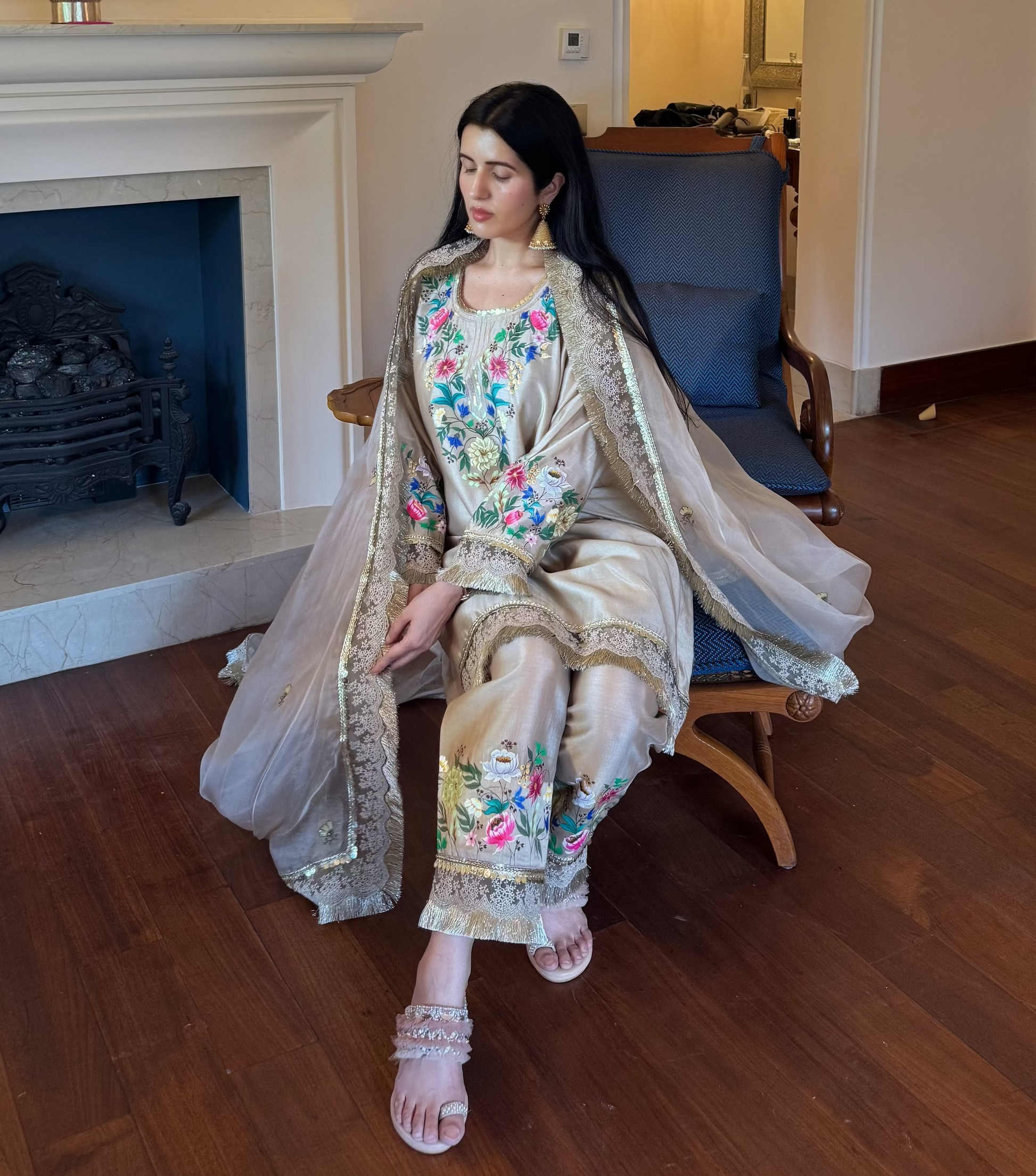 A woman wearing a traditional embroidered outfit sits on a blue armchair in a furnished room with a fireplace, wooden floor, and arched doorway.