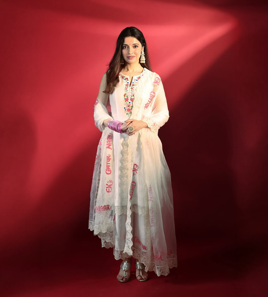A woman stands against a red studio backdrop wearing a long white embroidered traditional outfit with pink and green floral detailing, sheer matching fabric draped over her arm, and metallic silver shoes.

Divya Khosla IN Baavli