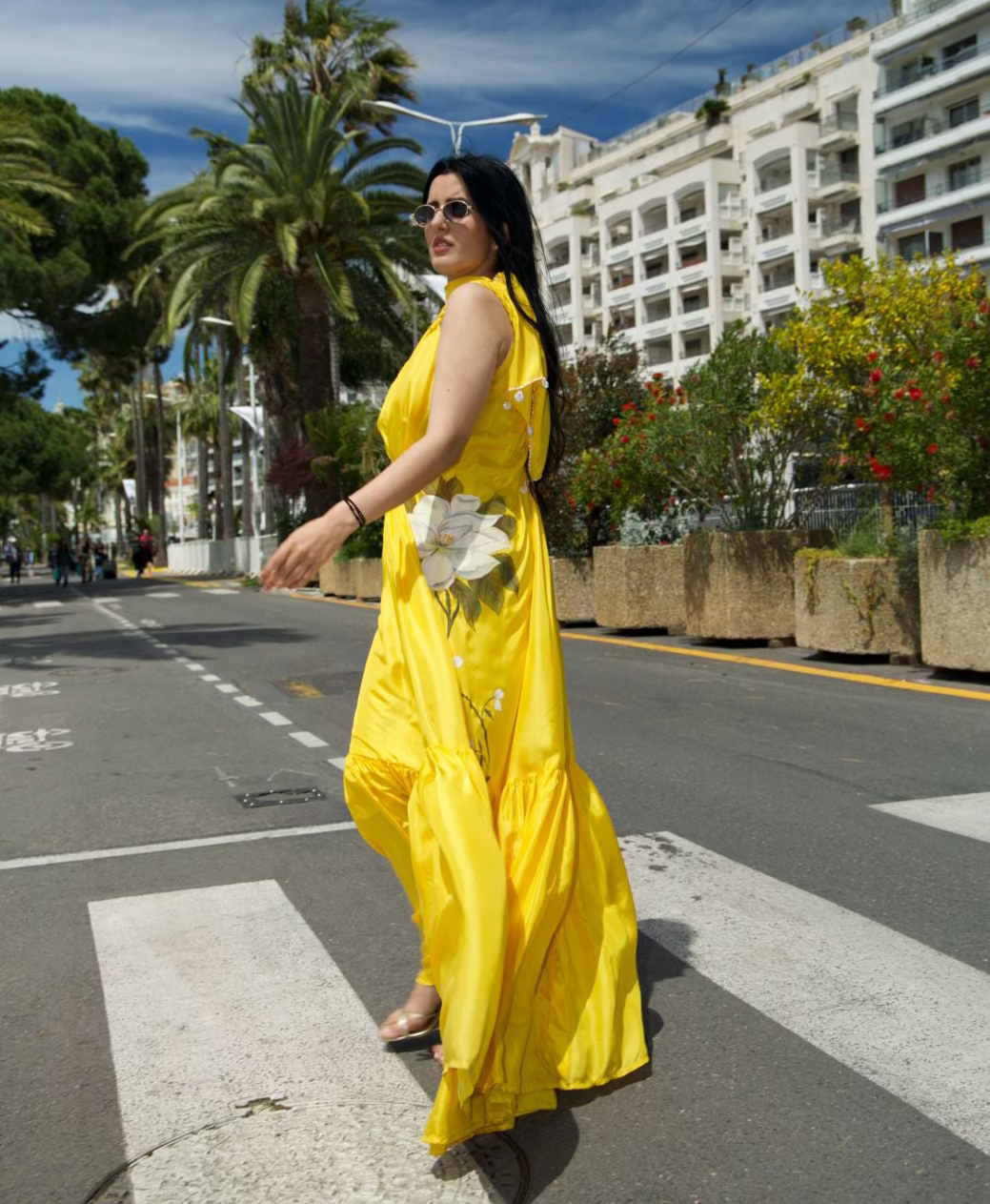 A woman wearing a bright yellow sleeveless maxi dress with white floral embroidery walks across a white-striped crosswalk on a city street. Palm trees, white buildings, and a blue sky are visible in the background.