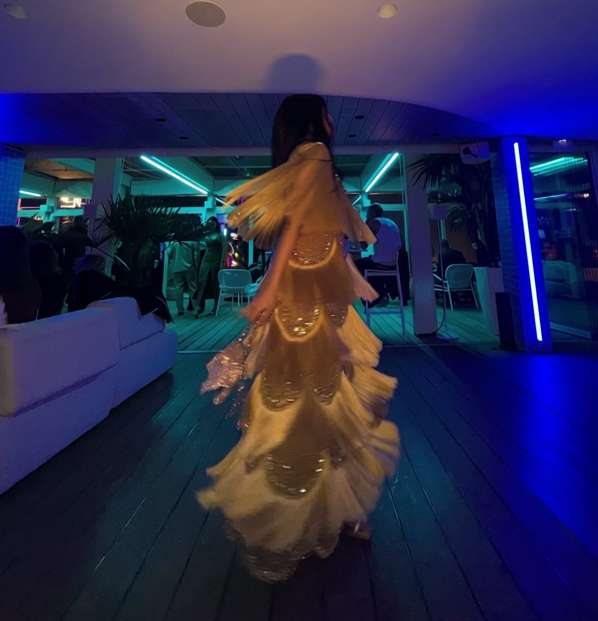 A woman in a flowing beige and gold embellished dress walks away in a dimly lit indoor venue with blue ambient lighting and a white sofa visible on the left.