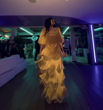 A woman wearing a shimmering gold tiered evening gown walks down a wooden hallway in a modern venue with purple ambient lighting.