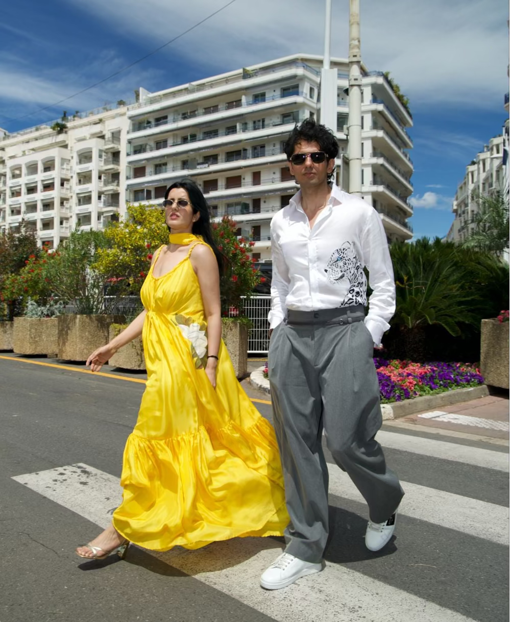A couple walking across a street crosswalk in front of white apartment buildings under a blue sky. The woman wears a bright yellow sleeveless gown, and the man wears a white long-sleeve shirt, gray trousers, and white sneakers.