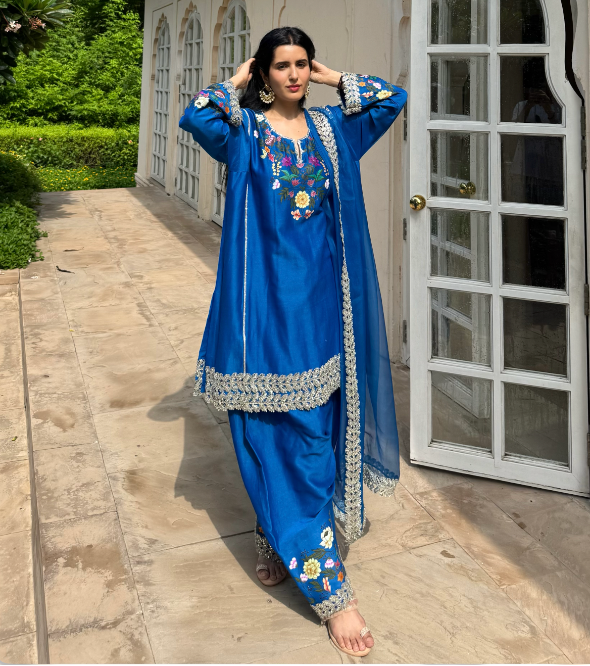 A woman models a royal blue traditional South Asian salwar kameez suit with floral embroidery and silver mirror work borders, standing on a stone patio in front of a beige building with white-framed glass doors.