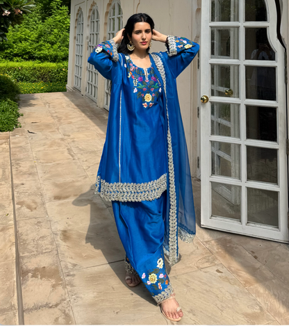 A woman models a royal blue traditional South Asian salwar kameez suit with floral embroidery and silver mirror work borders, standing on a stone patio in front of a beige building with white-framed glass doors.