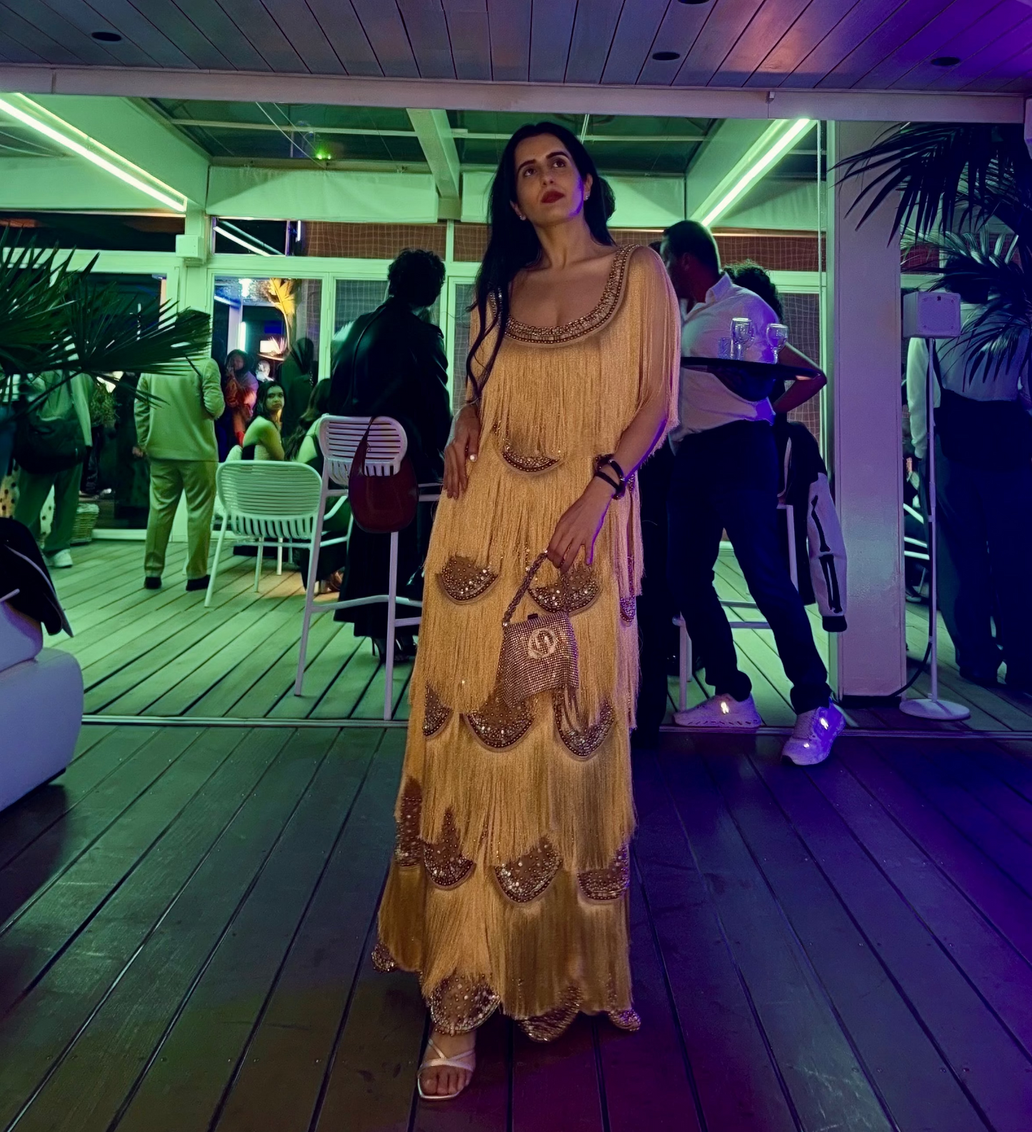 A woman wearing a long, flowing mustard yellow dress with embroidered details stands on a wooden deck in a modern indoor-outdoor venue with purple and green ambient lighting. Other people and plants are visible in the background.
