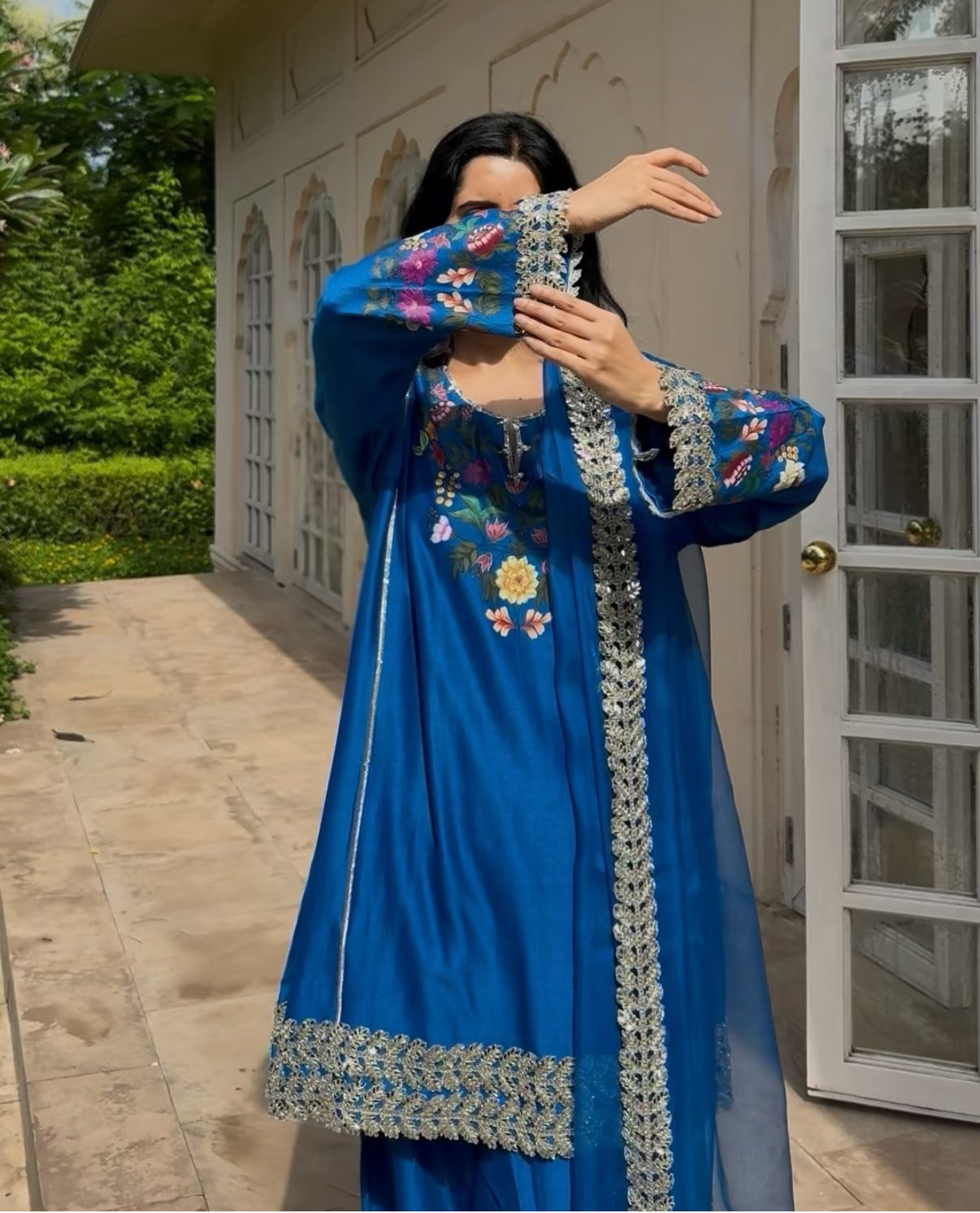 A woman wearing a vibrant blue traditional embroidered dress stands outdoors in front of a white building, holding up her hands in a pose that shows the sleeves and embroidery details.