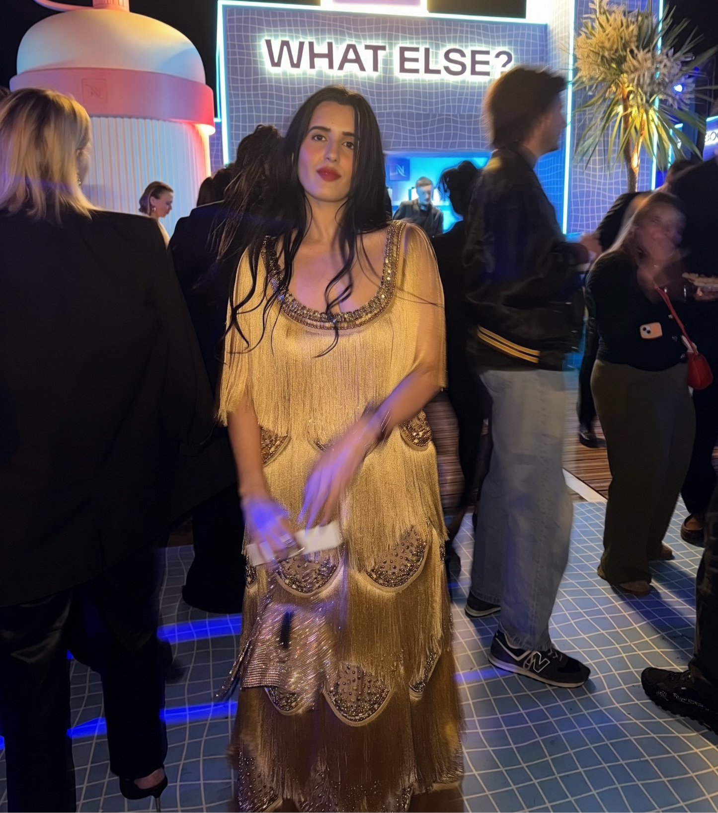 A woman wearing a gold fringed dress stands in a crowded event space at night, with a large illuminated sign reading 'WHAT ELSE?' and a stylized pink cup decoration behind her.