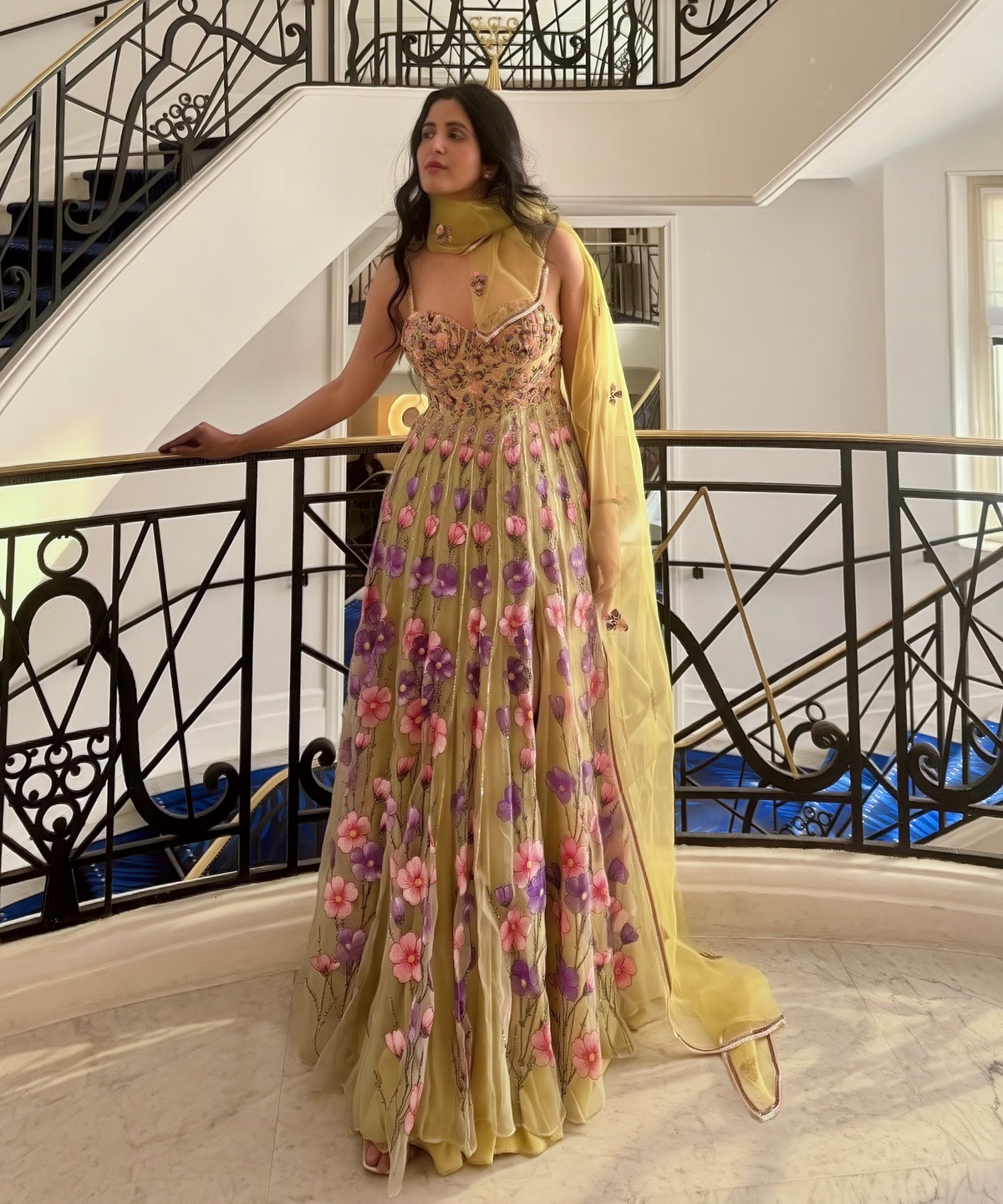 A woman models a floor-length yellow embellished gown with a matching dupatta in an elegant indoor setting with a staircase.