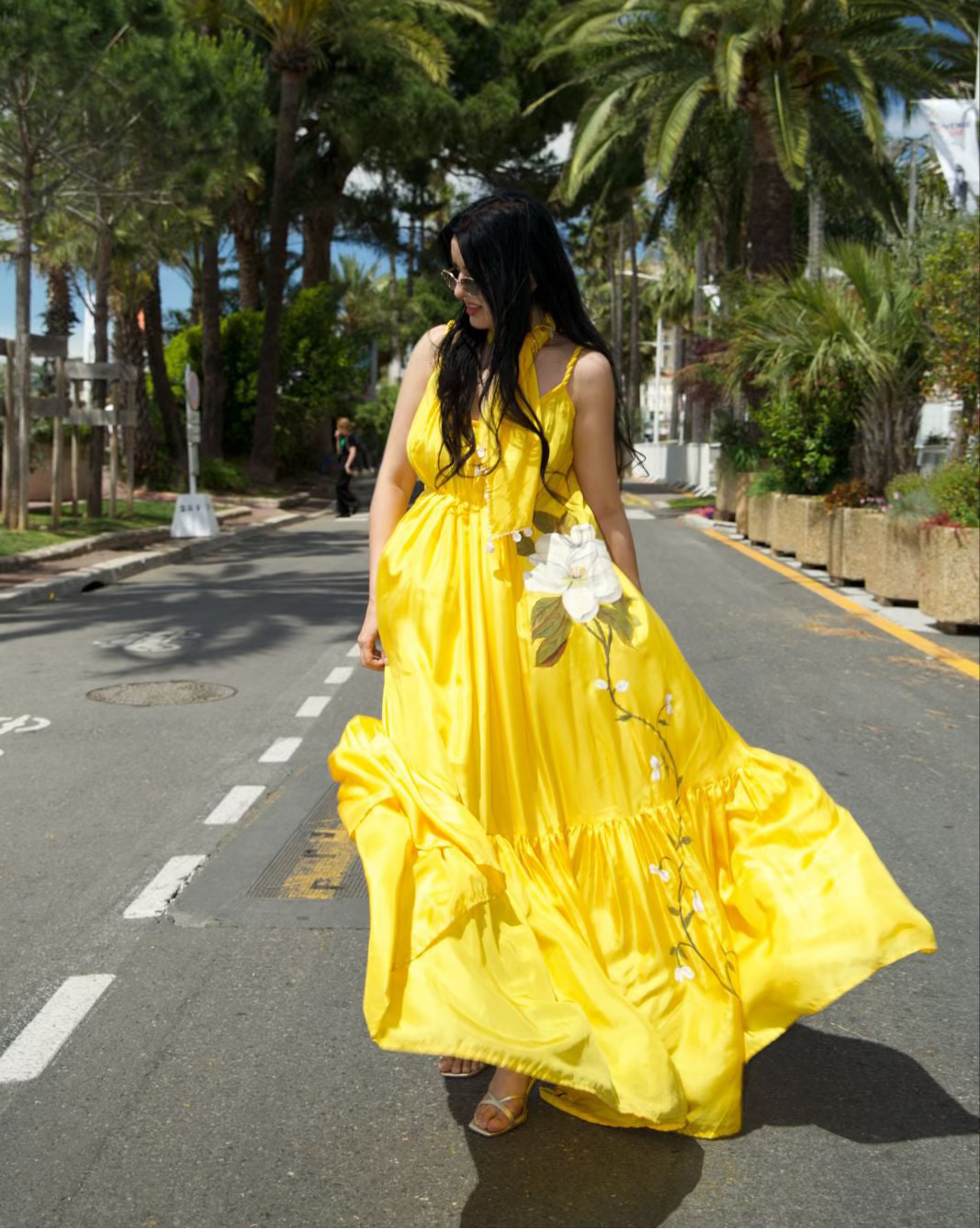 A woman walking on a palm-tree-lined street wearing a bright yellow sleeveless maxi dress with white floral embroidery and holding a bouquet of white flowers.