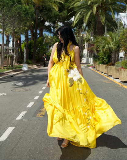 A woman walking on a palm-tree-lined street wearing a bright yellow sleeveless maxi dress with white floral embroidery and holding a bouquet of white flowers.