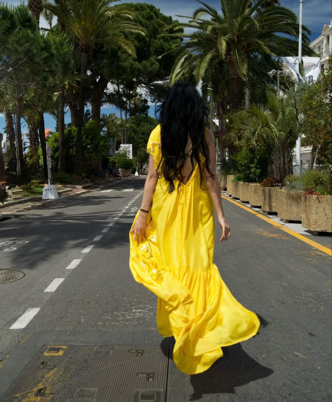 A woman with long black hair is seen from behind, wearing a flowing bright yellow maxi dress while walking down a palm-tree-lined street.