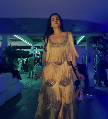 A woman wearing a beige fringed maxi dress with metallic embellishments, standing in a dimly lit event space with blue ambient lighting.