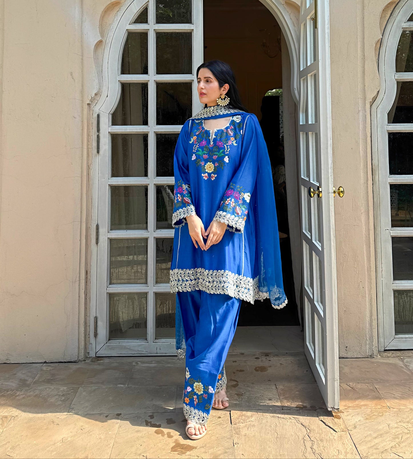 A woman wearing a vibrant blue embroidered traditional South Asian outfit with a matching dupatta stands in an open doorway of a beige building.