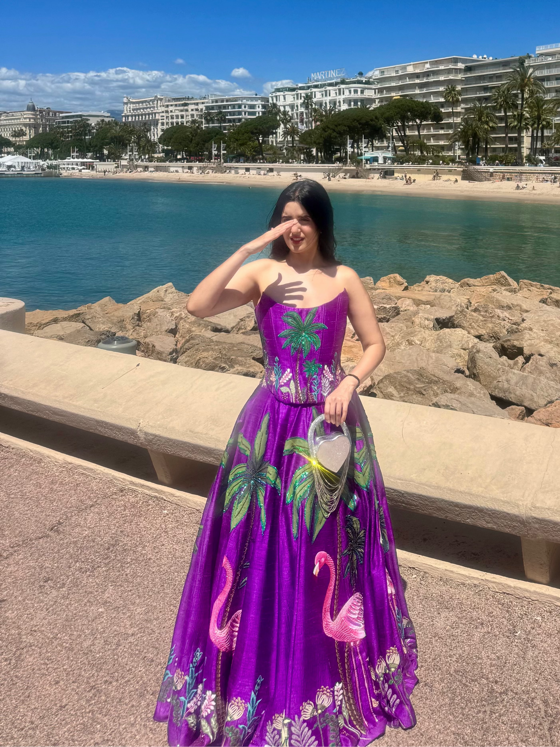 A woman wearing a vibrant purple and green tropical flamingo print maxi dress stands on a seaside promenade with a beach, ocean, and coastal buildings in the background.