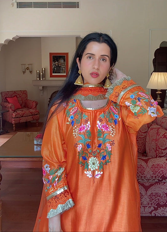 A woman wearing a vibrant orange embroidered kurta with colorful floral patterns and gold jewelry, standing in a decorated living room with a chandelier and sofas in the background.