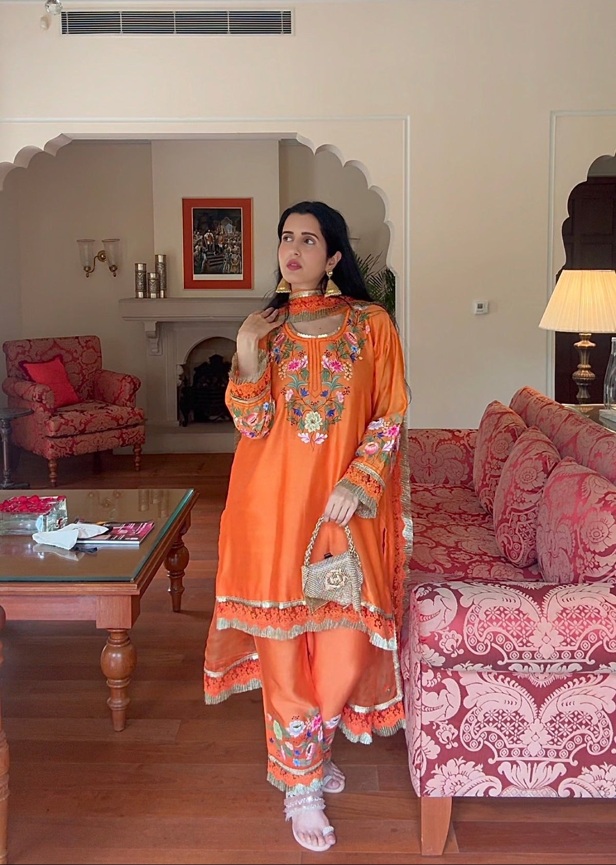 A woman modeling a bright orange traditional South Asian outfit with intricate multicolor embroidery, standing in a decorated living room with pink patterned sofas and wooden floors.