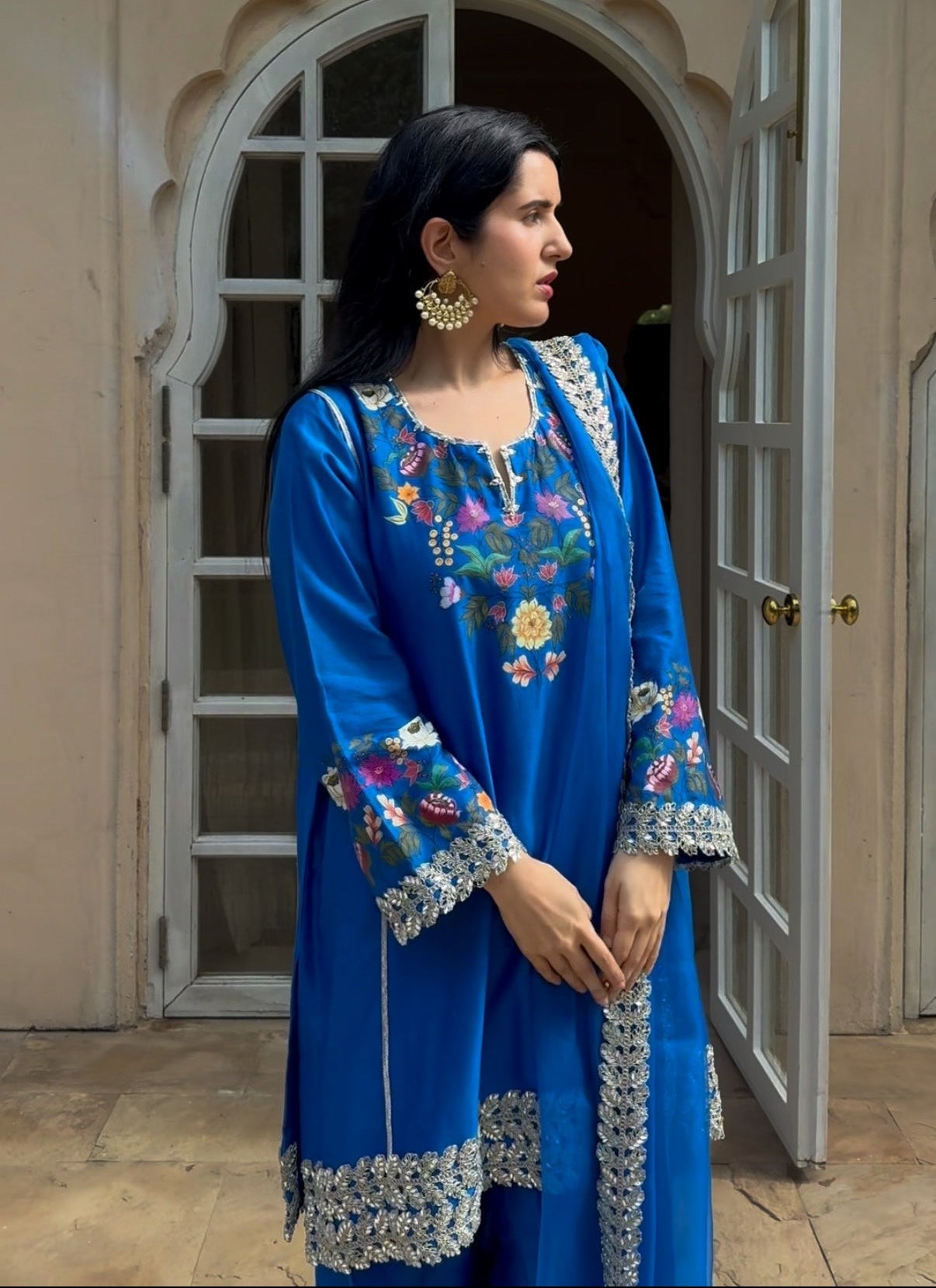 A woman wearing a royal blue embroidered kurta with floral patterns and silver-toned lace trim, standing in front of an arched doorway.