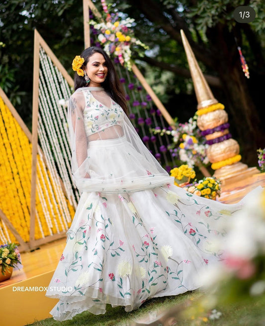 A woman models a white floral embroidered lehenga choli set outdoors on a decorated stage with yellow garlands and green foliage in the background.