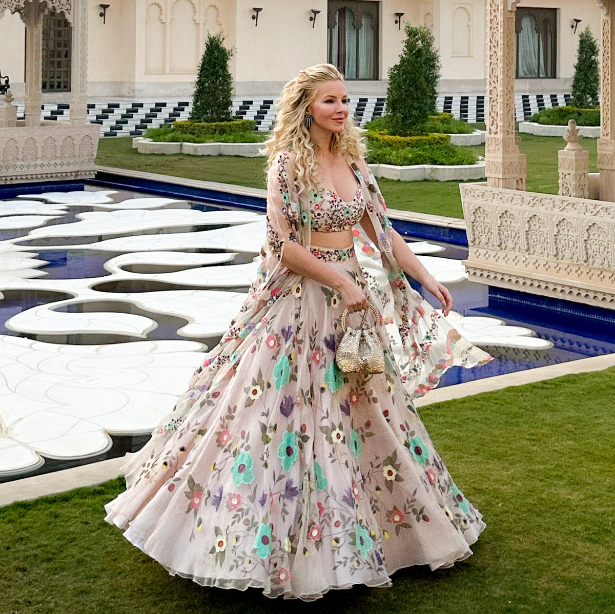 A woman wearing a light pink floral lehenga choli traditional Indian outfit stands in front of a grand palace courtyard with a reflecting pool.