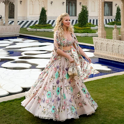 A woman wearing a light pink floral lehenga choli traditional Indian outfit stands in front of a grand palace courtyard with a reflecting pool.