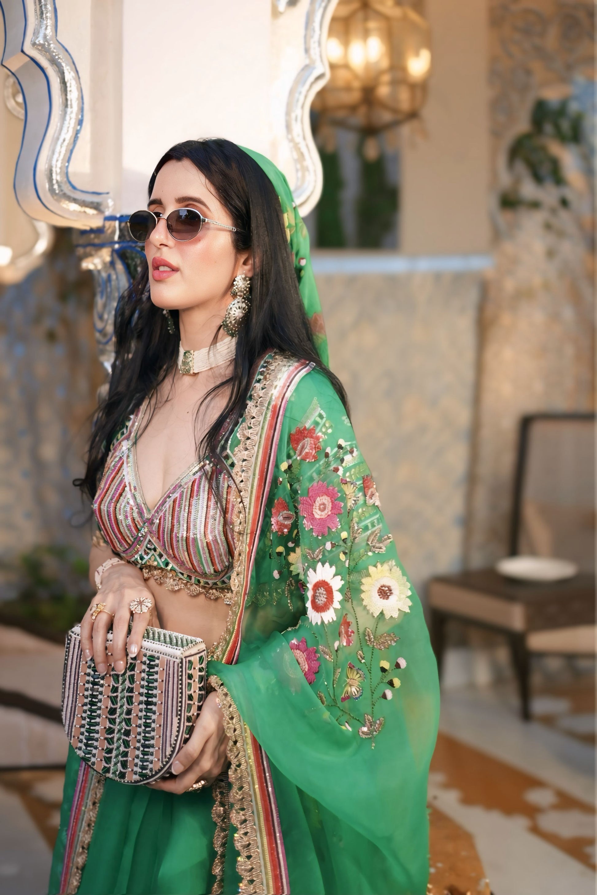 A woman wearing a vibrant green embroidered traditional outfit with a matching headscarf, sunglasses, and a patterned clutch, posed in an elegant indoor setting with a mirror and warm lighting.