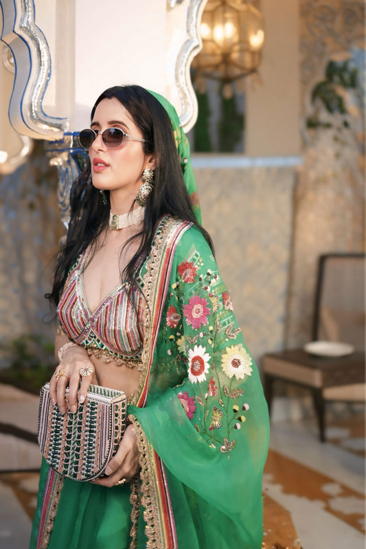 A woman wearing a vibrant green embroidered traditional outfit with a matching headscarf, sunglasses, and a patterned clutch, posed in an elegant indoor setting with a mirror and warm lighting.