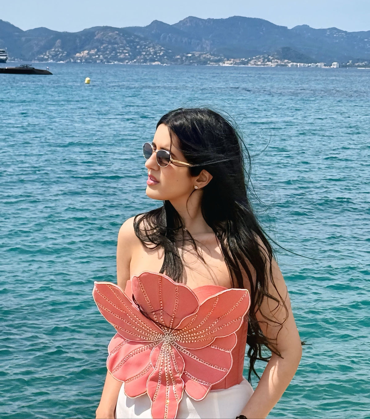 A woman with long dark hair and sunglasses stands by the sea wearing a strapless coral-pink top with a large flower-shaped embellishment on the front, with blue ocean water and mountains in the background.
