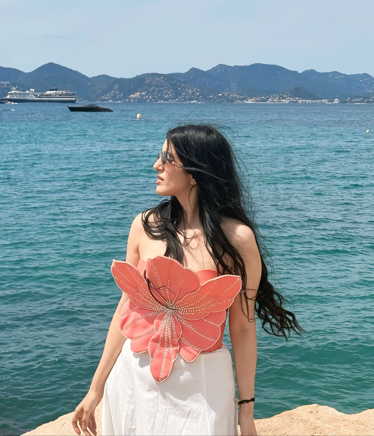 A woman with long black hair stands on a rocky shoreline with a turquoise sea and distant mountains behind her. She is wearing a white dress with a large coral-pink flower applique on the front.
