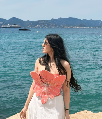 A woman with long black hair stands on a rocky shoreline with a turquoise sea and distant mountains behind her. She is wearing a white dress with a large coral-pink flower applique on the front.