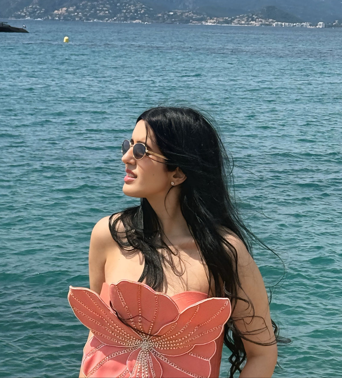 A woman with long black hair and sunglasses stands by the sea wearing a coral-pink strapless dress with a large flower-like petal detail on the front, with blue ocean water and mountains in the background.