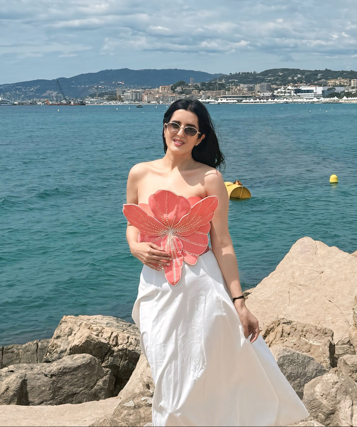 A woman wearing a white strapless maxi dress and sunglasses stands on rocks by the sea, holding a large pink flower-shaped prop. The background features a scenic coastal view with blue water, distant hills, and a cloudy sky.